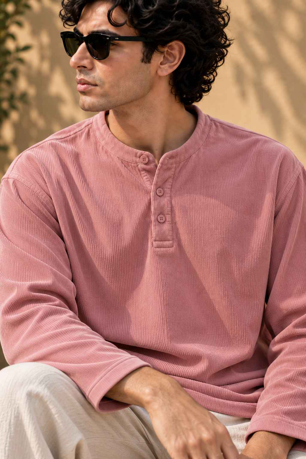 A man wearing a dusty rose pink corduroy long-sleeve henley shirt and beige trousers, sitting outdoors against a warm beige wall with soft shadows.