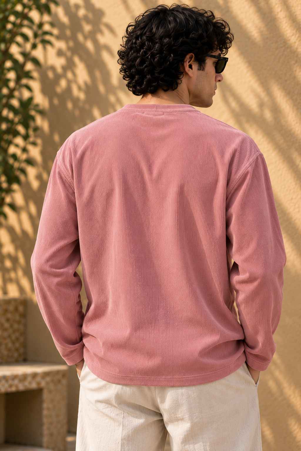 A man viewed from behind wearing a dusty rose pink long-sleeve crewneck shirt and off-white trousers, standing outdoors against a textured beige wall with leaf shadows.