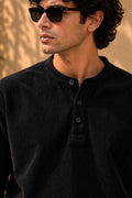 A man wearing a black corduroy henley shirt and black sunglasses, shown in a close-up profile shot against a warm, sunlit beige background.