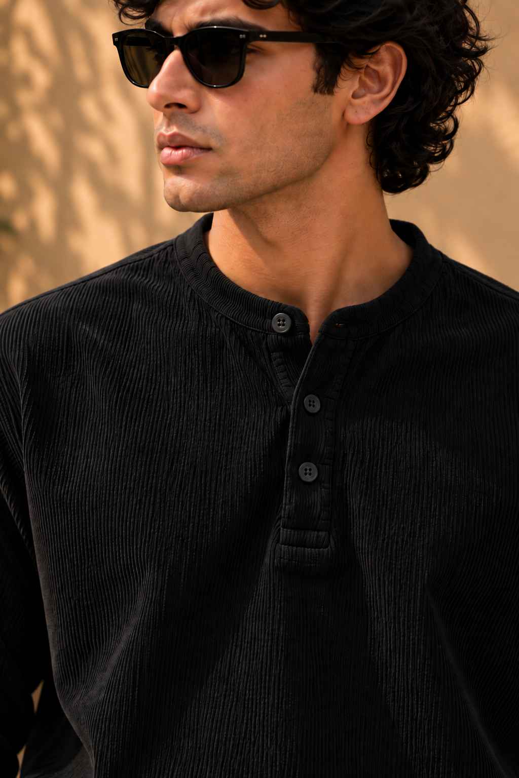 A man wearing a black corduroy henley shirt and black sunglasses, shown in a close-up profile shot against a warm, sunlit beige background.