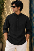 A man wearing a black long-sleeve mandarin collar henley shirt with white trousers, standing outdoors against a textured beige wall.