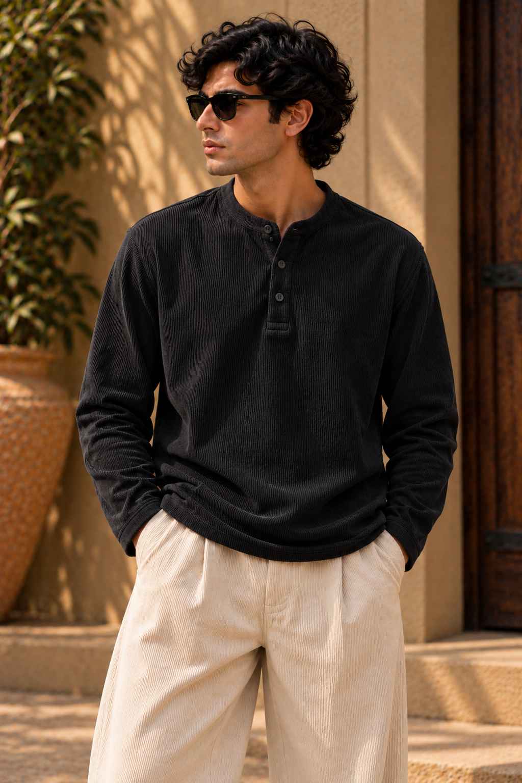 A man wearing a black long-sleeve henley shirt and cream-colored trousers, standing outdoors in front of a beige wall and wooden door.