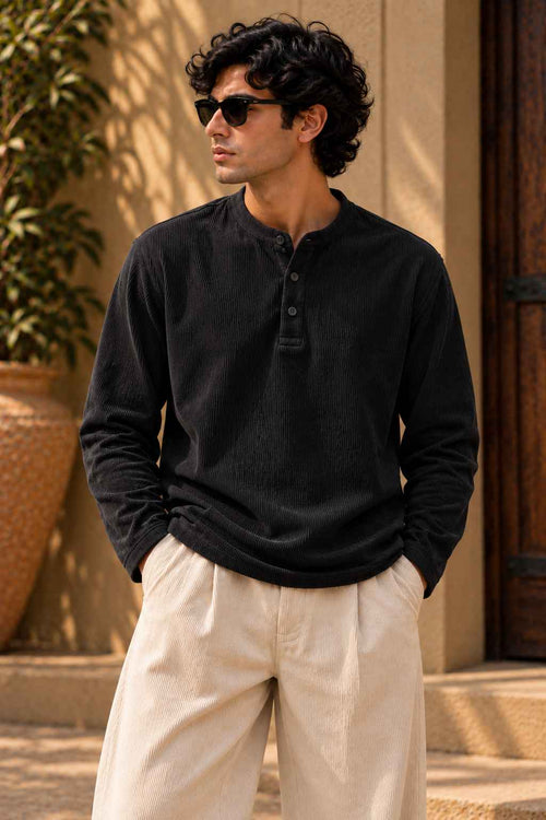 A man wearing a black long-sleeve henley shirt and cream-colored trousers, standing outdoors in front of a beige wall and wooden door.