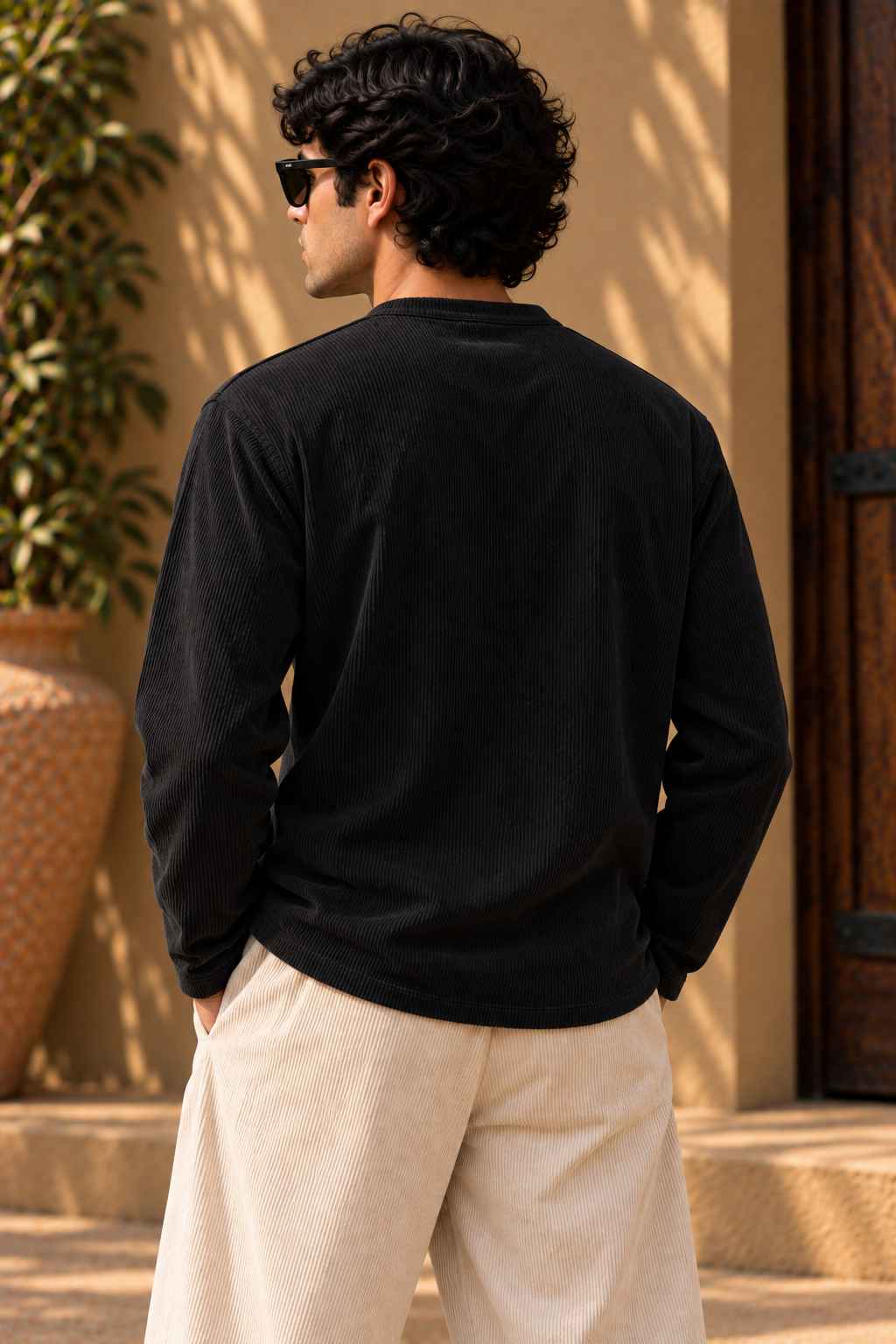 A man wearing a black long-sleeve crewneck shirt and cream-colored pants, photographed from behind in a warm, sunlit outdoor setting with a beige stucco wall and potted plants.