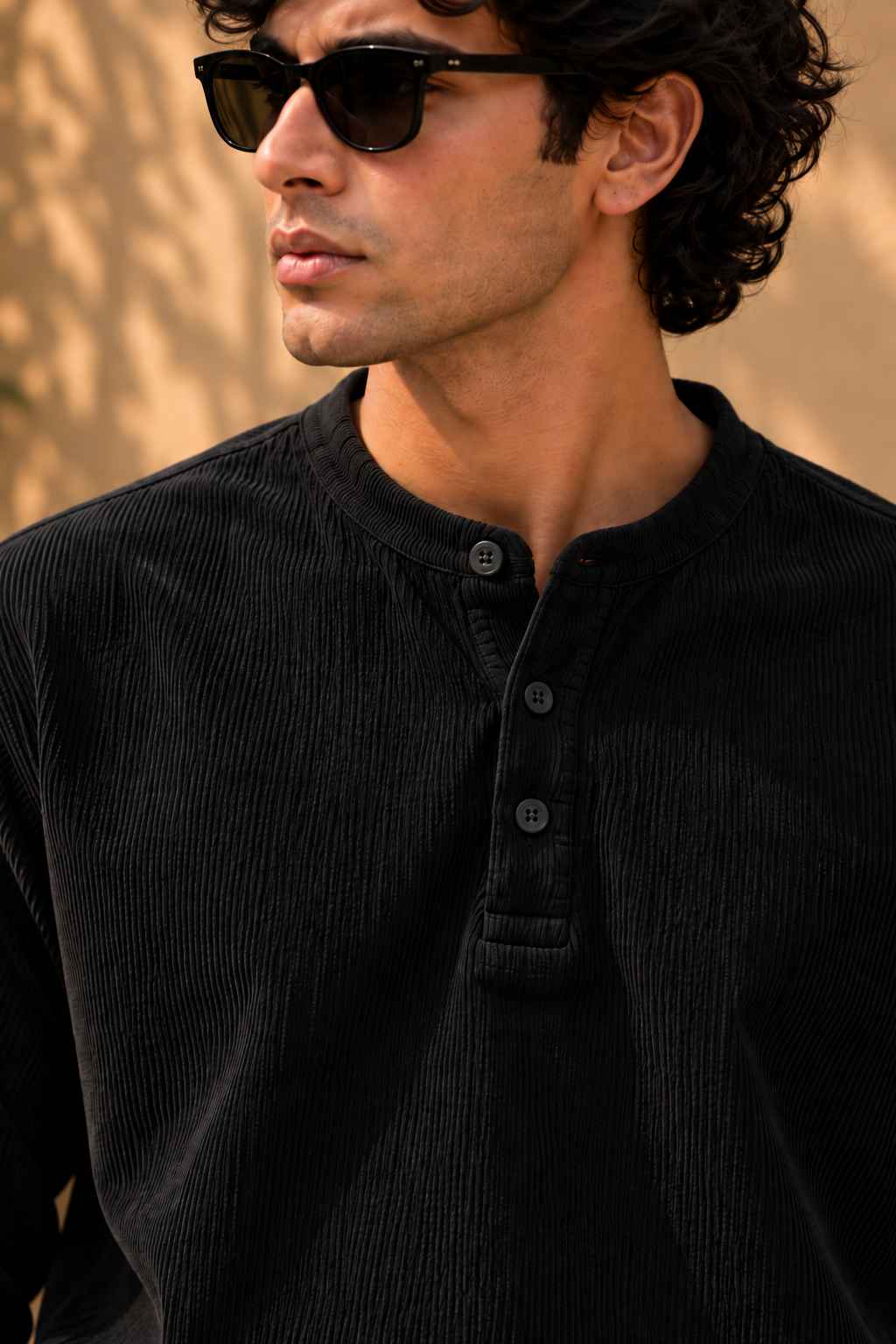 A man wearing black sunglasses and a black corduroy henley shirt, shown in a close-up profile against a warm beige background.