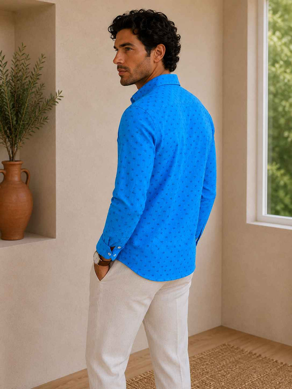 Man wearing a blue textured shirt and beige pants standing in a room with a plant and window.