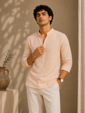Man wearing a light pink shirt and white pants standing against a beige wall with a plant in the background.