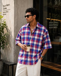 Man wearing a plaid shirt and sunglasses, holding a coffee cup, standing outside a cafe.