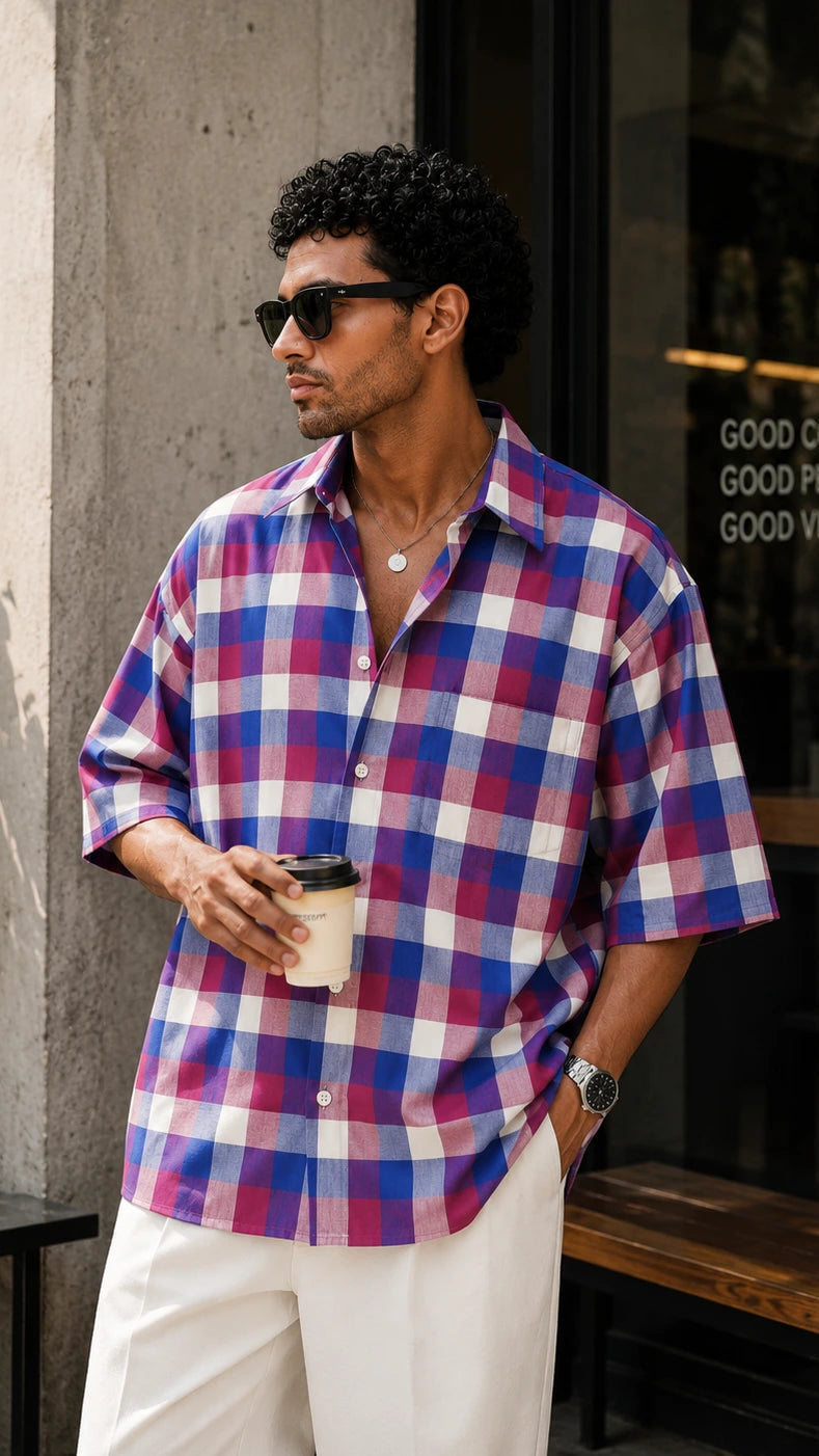 Man wearing a plaid shirt and sunglasses, holding a coffee cup, standing outside a cafe.