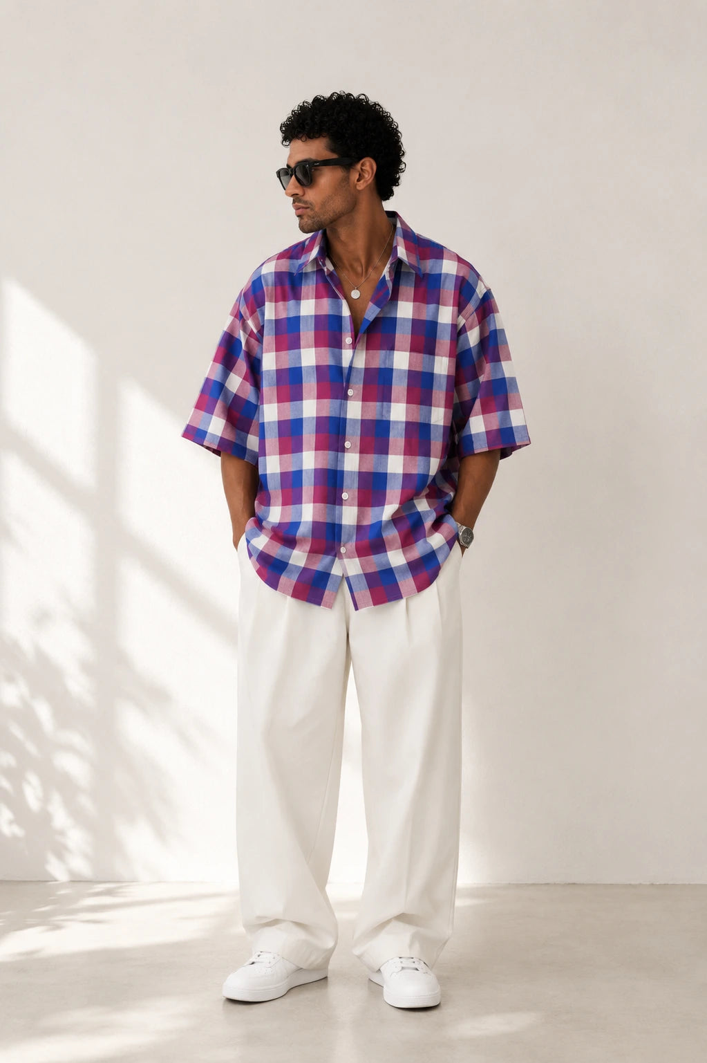 Man wearing a red, blue, and white checkered shirt with white pants standing against a white wall.