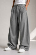 A person wearing very wide-leg pleated gray trousers, styled with a white t-shirt and white sneakers, standing against a neutral studio background.
