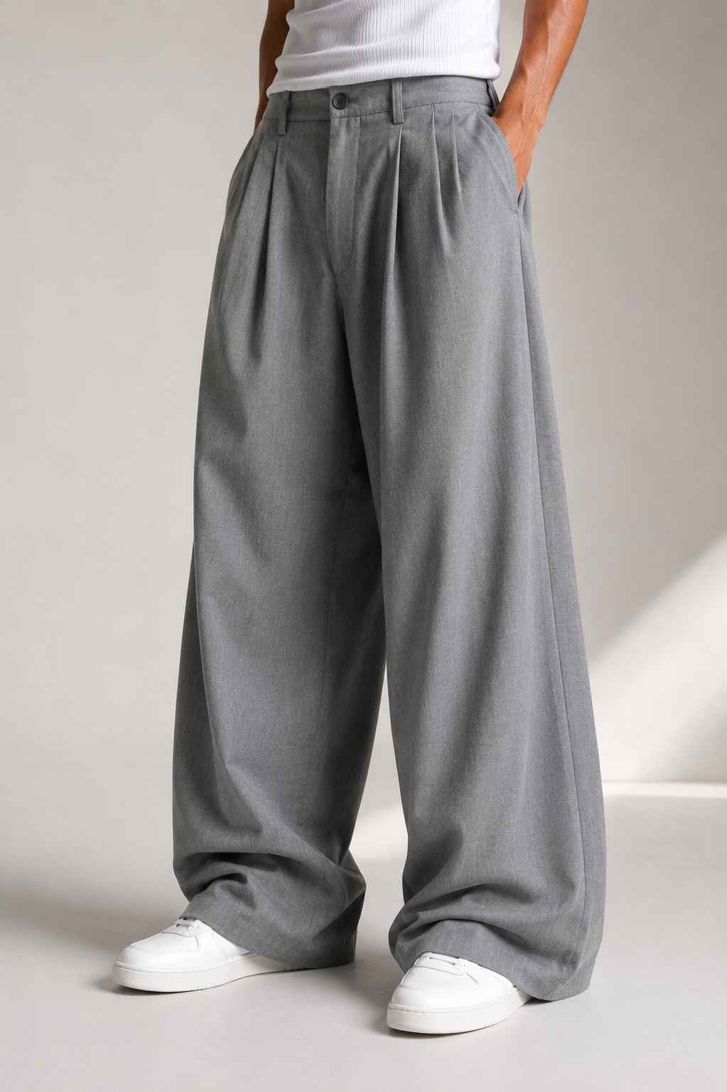 A person wearing wide-leg gray pleated trousers with a white top and white sneakers, photographed in a studio setting.
