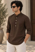 A man wearing a dark brown long-sleeve kurta with a mandarin collar and white pants, standing in a sunlit outdoor setting.