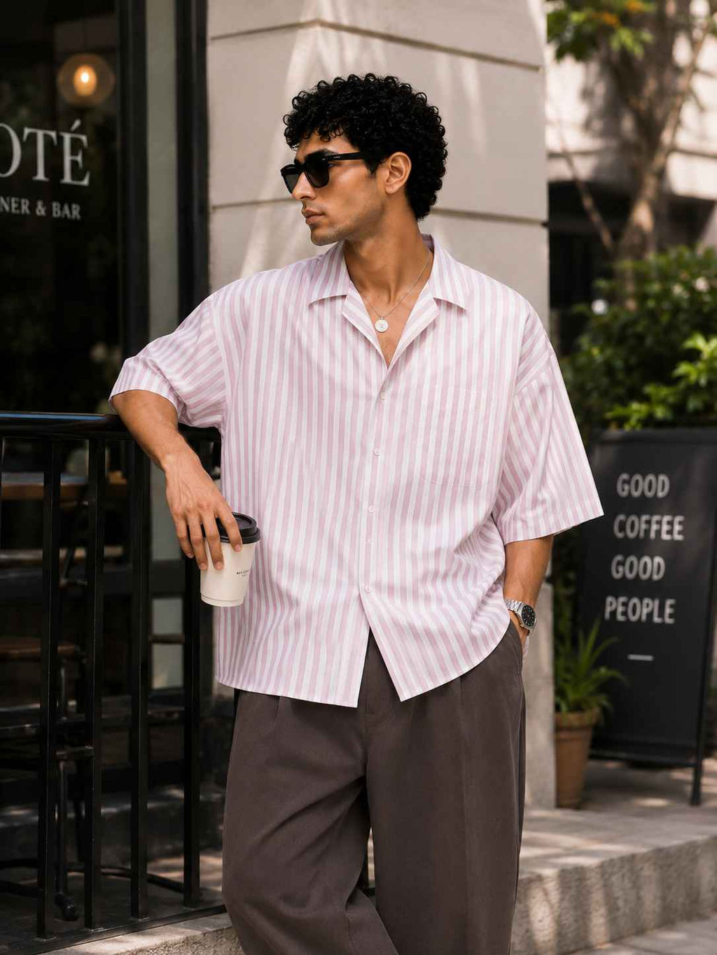 Man in a striped shirt and sunglasses holding a coffee cup, standing on a street.