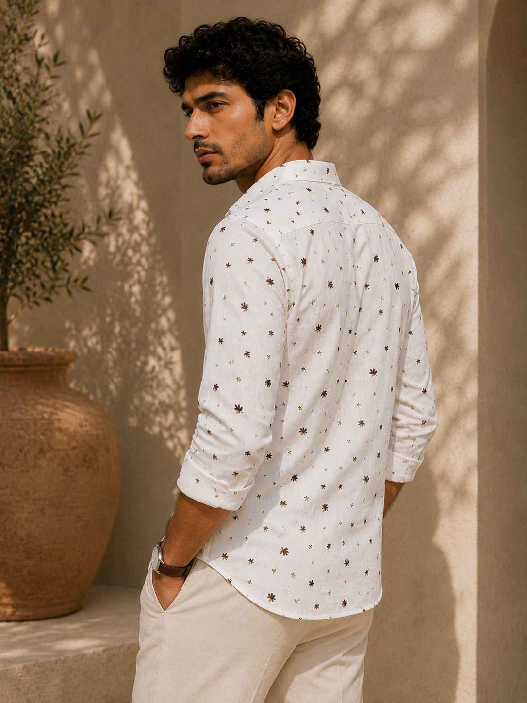 Man wearing a white patterned shirt standing against a beige wall with a potted plant.