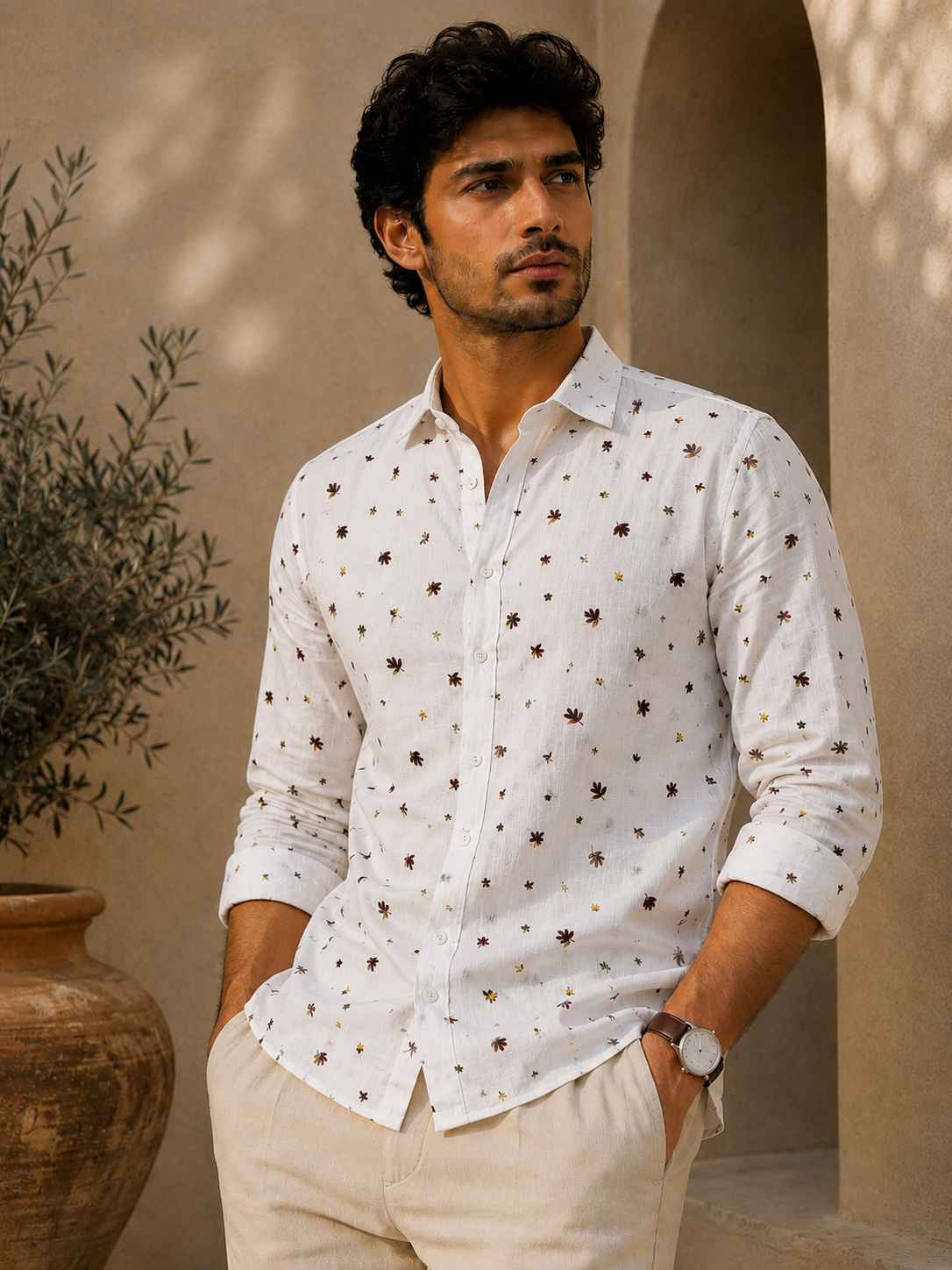 Man wearing a white patterned shirt standing against a beige wall with a plant in the background