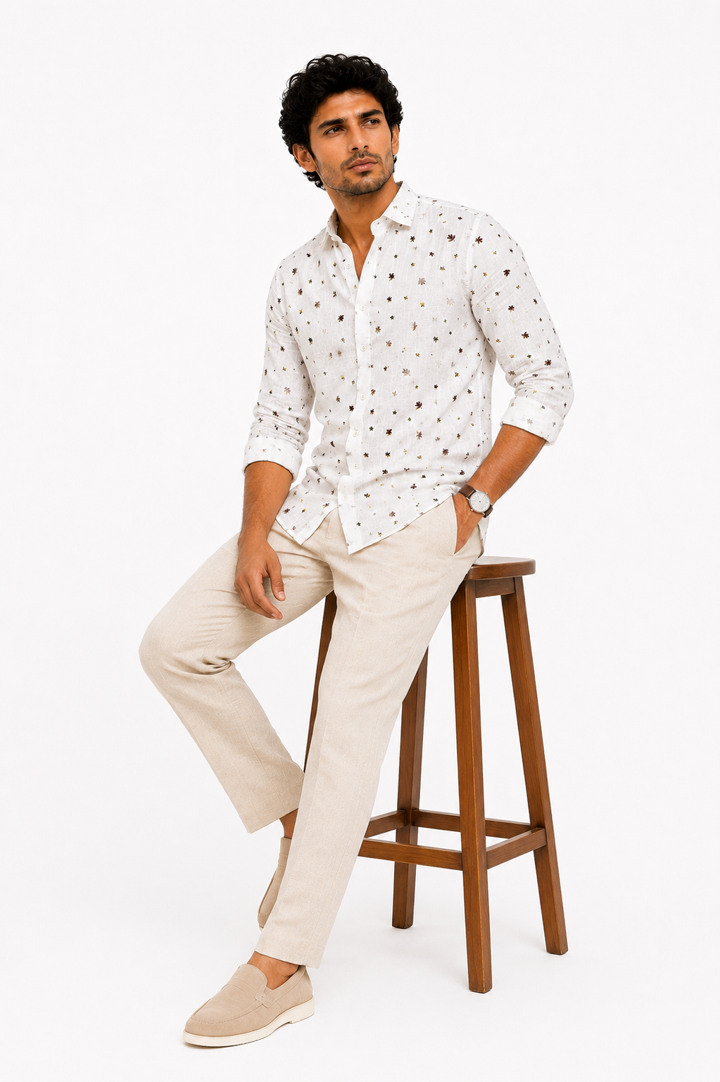 Man wearing a white patterned shirt and beige pants sitting on a wooden stool against a white background
