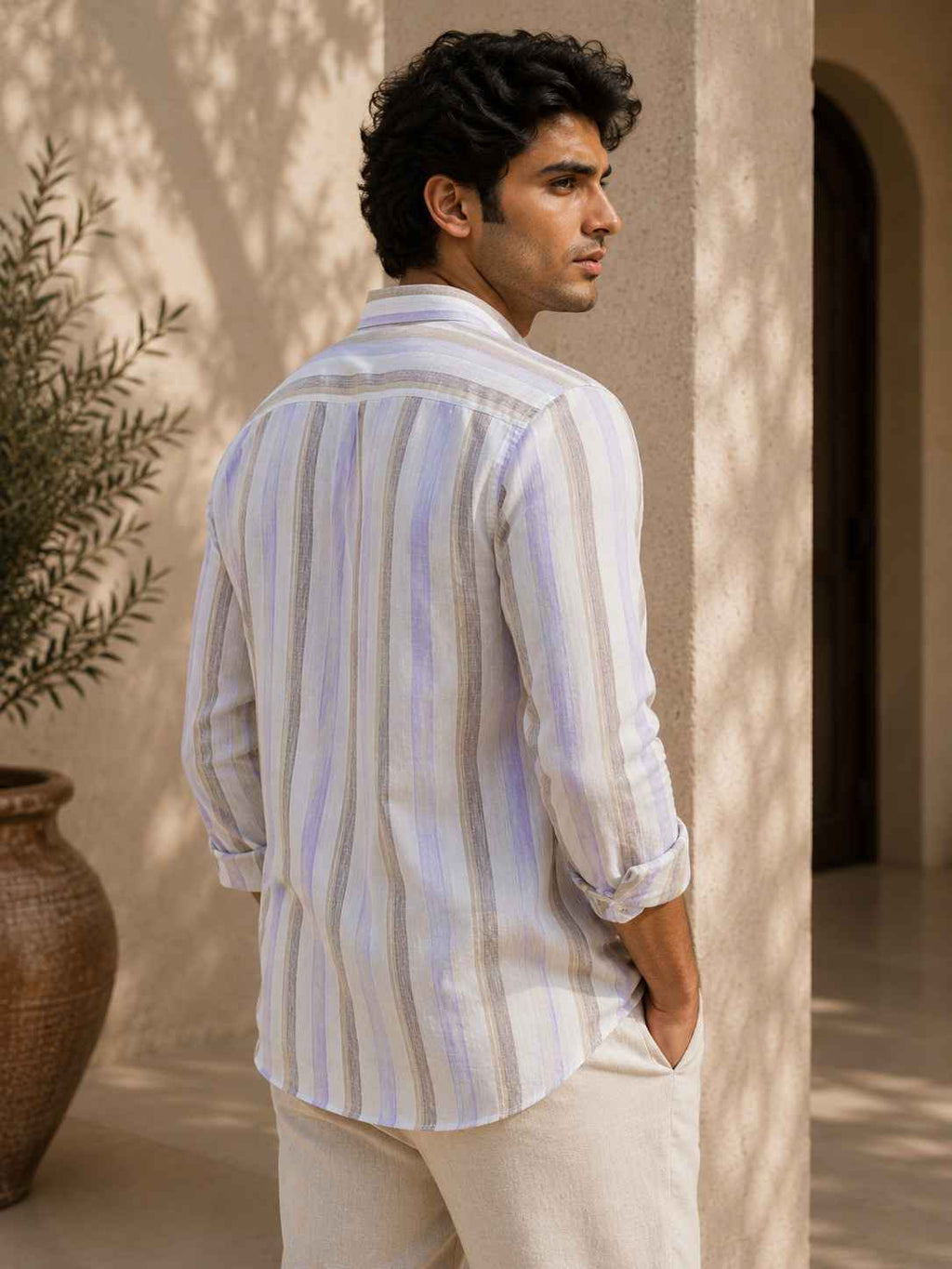 Man wearing a striped shirt and white pants standing against a beige wall with a plant in the background