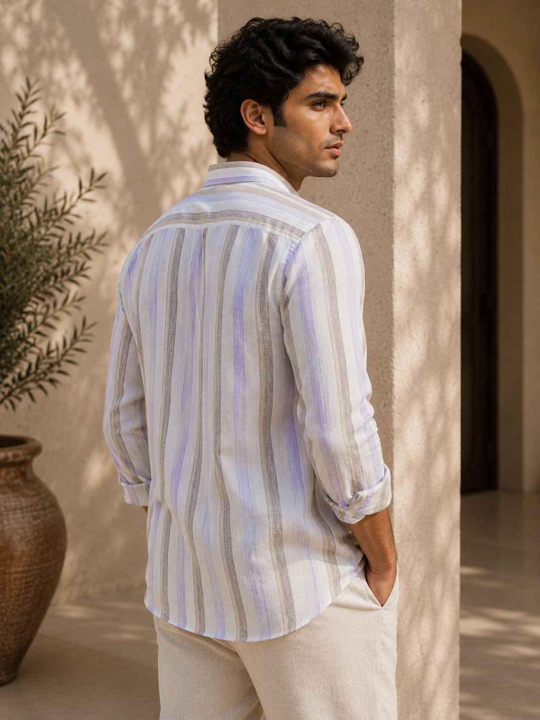 Man wearing a striped shirt and white pants standing against a beige wall with a plant in the background