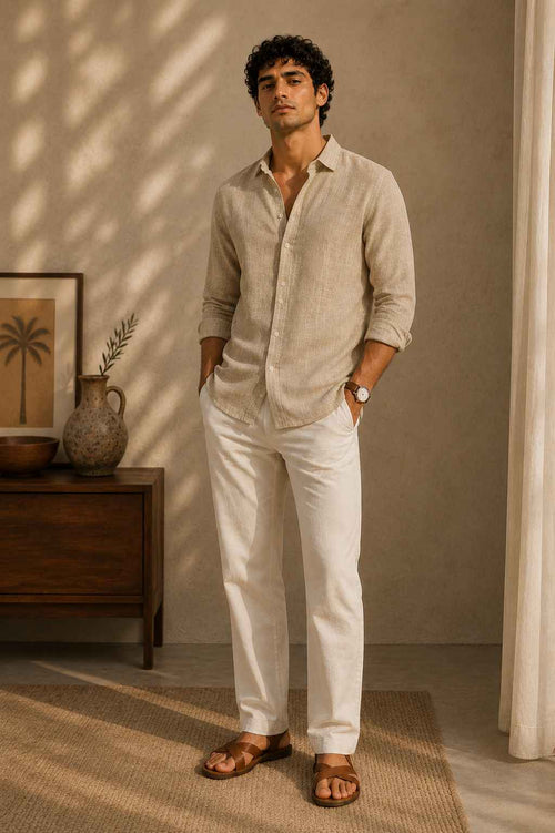 Man wearing a beige shirt and white pants standing in a room with a wooden side table and decorative items.
