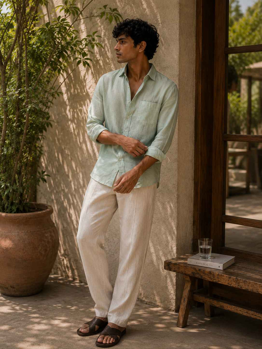 A man wearing a light sage green linen button-down shirt and off-white linen trousers, standing on a patio next to a wooden bench and potted plants.