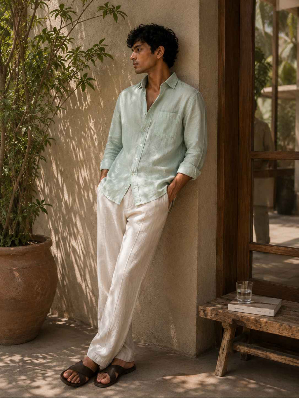 A man wearing a light mint green long-sleeve button-up shirt and off-white linen trousers, leaning against a stucco wall next to a potted plant.