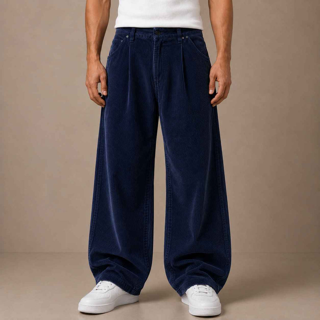 A person wearing very wide-leg navy corduroy pants with a white t-shirt and white sneakers, standing against a plain beige studio background.