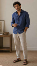 Man wearing a blue shirt and beige pants standing in a room with a wooden shelf and plant.