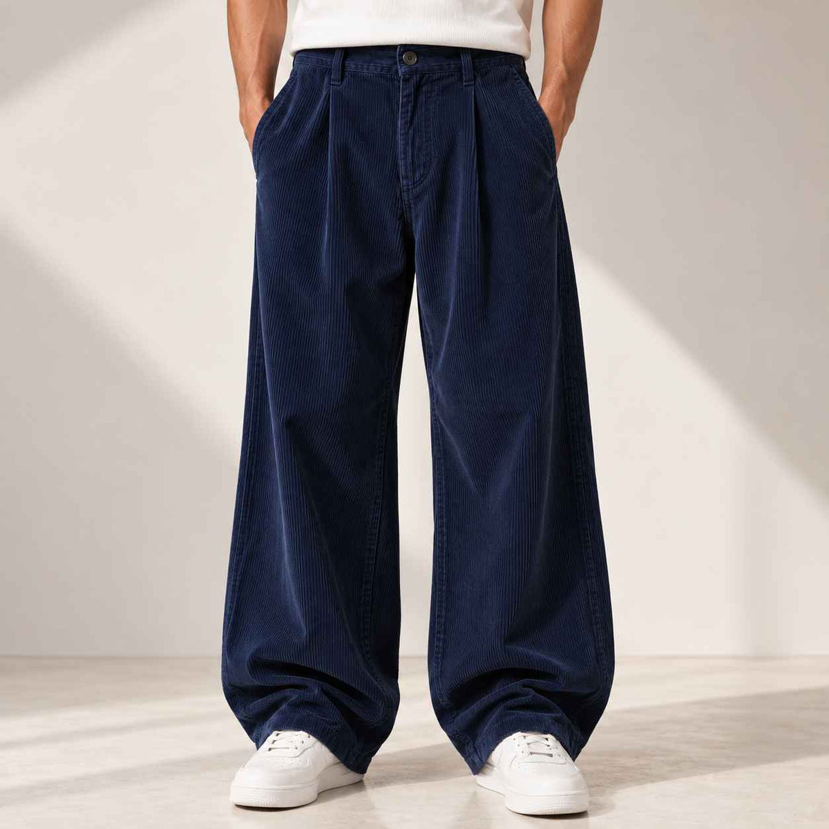 A person wearing very wide-leg navy corduroy pants with a pleated front, paired with a white t-shirt and white sneakers, standing against a minimalist beige wall with soft diagonal shadows.