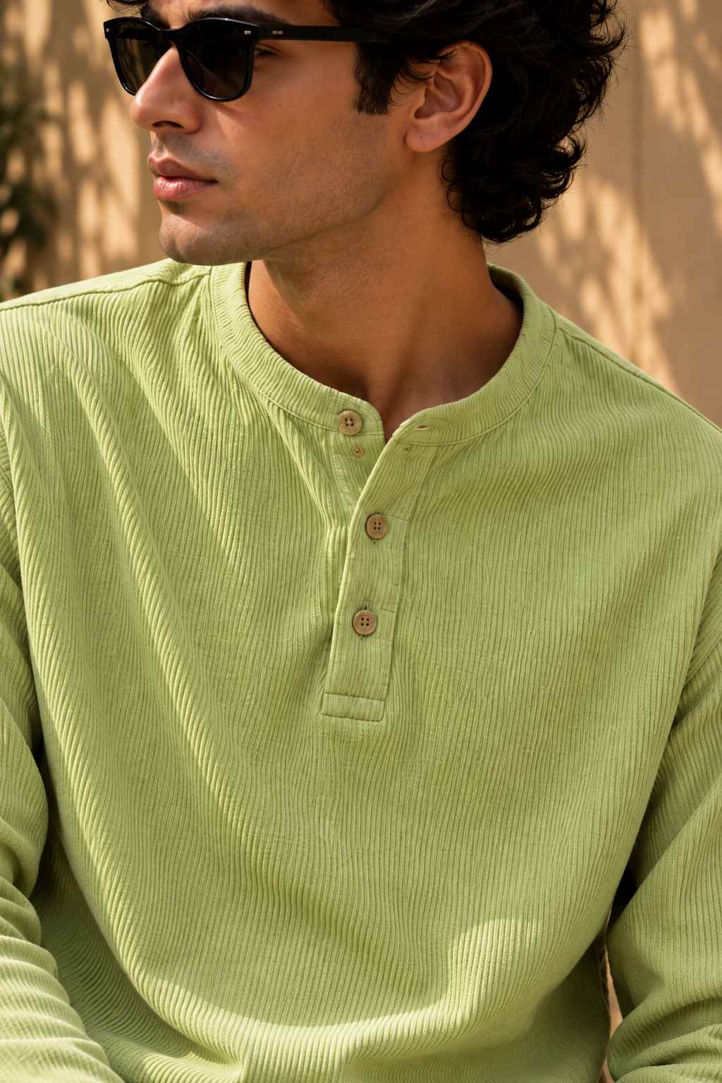 A man wearing a light sage green corduroy henley shirt with three buttons, styled with black sunglasses against a warm, sunlit background.