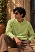 A man wearing a light green long-sleeve henley shirt and brown pants sits outdoors on stone steps, wearing dark sunglasses.