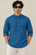 A man wearing a blue long-sleeve mandarin collar henley shirt with white buttons, paired with off-white trousers and black sunglasses, standing against a plain beige background.