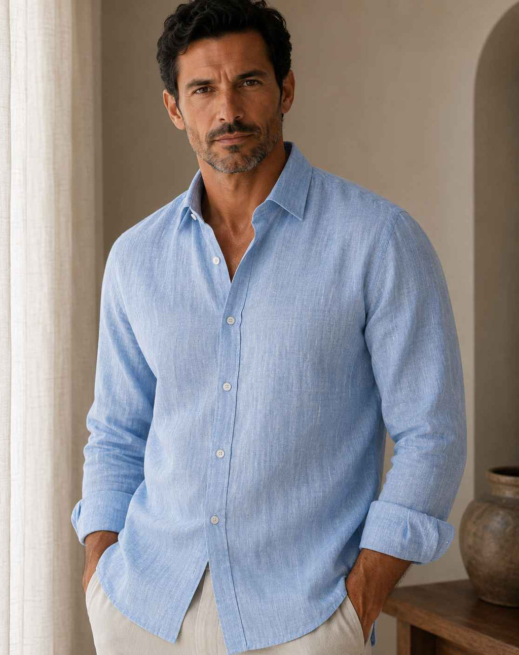 Man wearing a light blue shirt in a room with a vase and curtain.