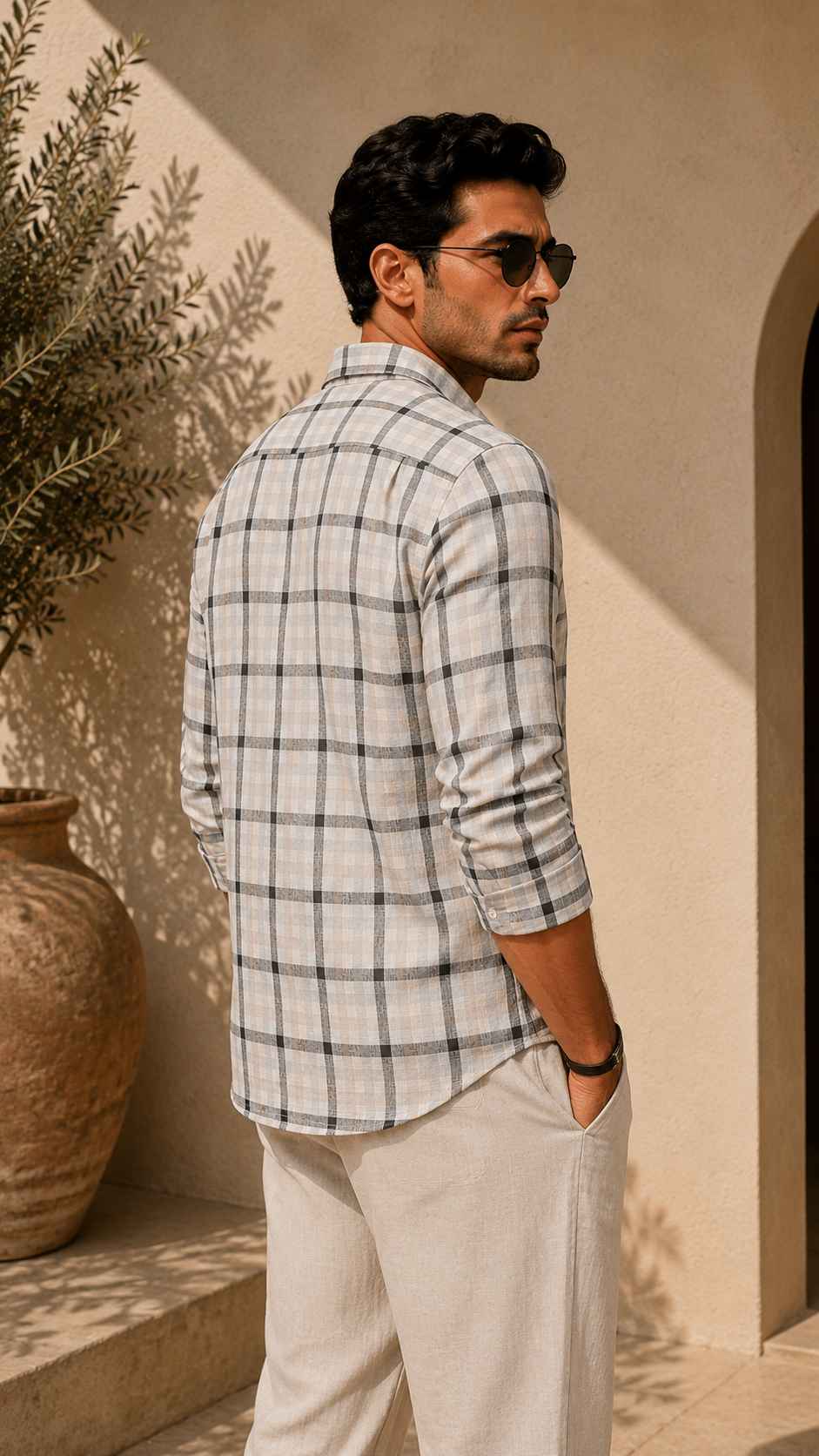 Man wearing a checkered shirt and sunglasses standing against a beige wall.