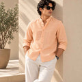 A man wearing a light peach linen long-sleeve button-up shirt and white trousers, leaning against a beige wall in a sunlit outdoor setting with a potted plant in the background.
