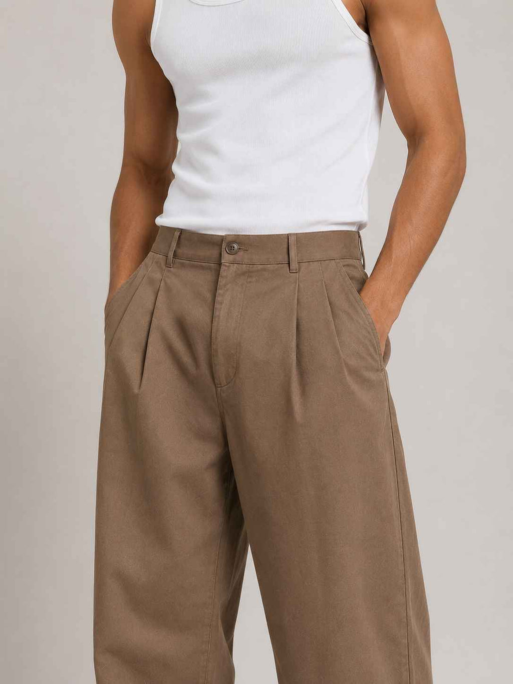 A person wearing pleated wide-leg brown trousers and a white ribbed tank top, standing against a plain light gray studio background.