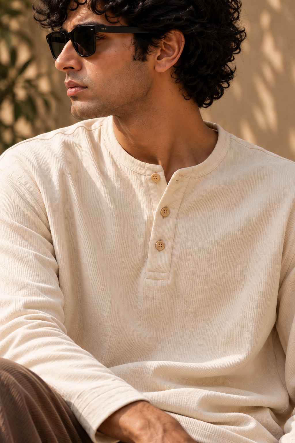 A man wearing a cream-colored ribbed long-sleeve henley shirt and black sunglasses, captured in a warm, sunlit outdoor setting.