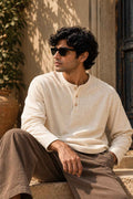 A man wearing a cream-colored long-sleeve henley shirt and brown trousers sits on stone steps outdoors, wearing dark sunglasses.