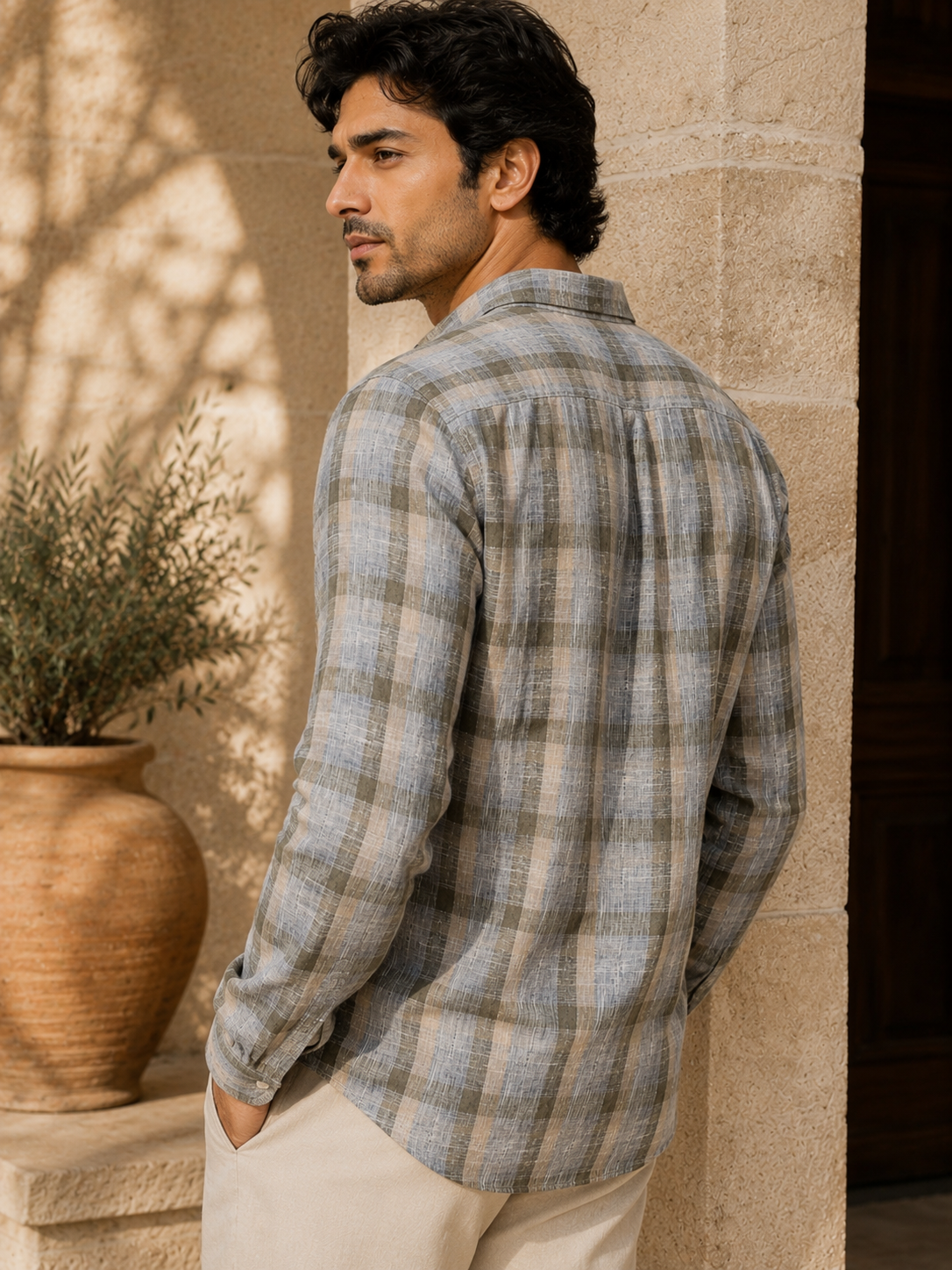 Man wearing a plaid shirt standing against a stone wall with a plant in the background