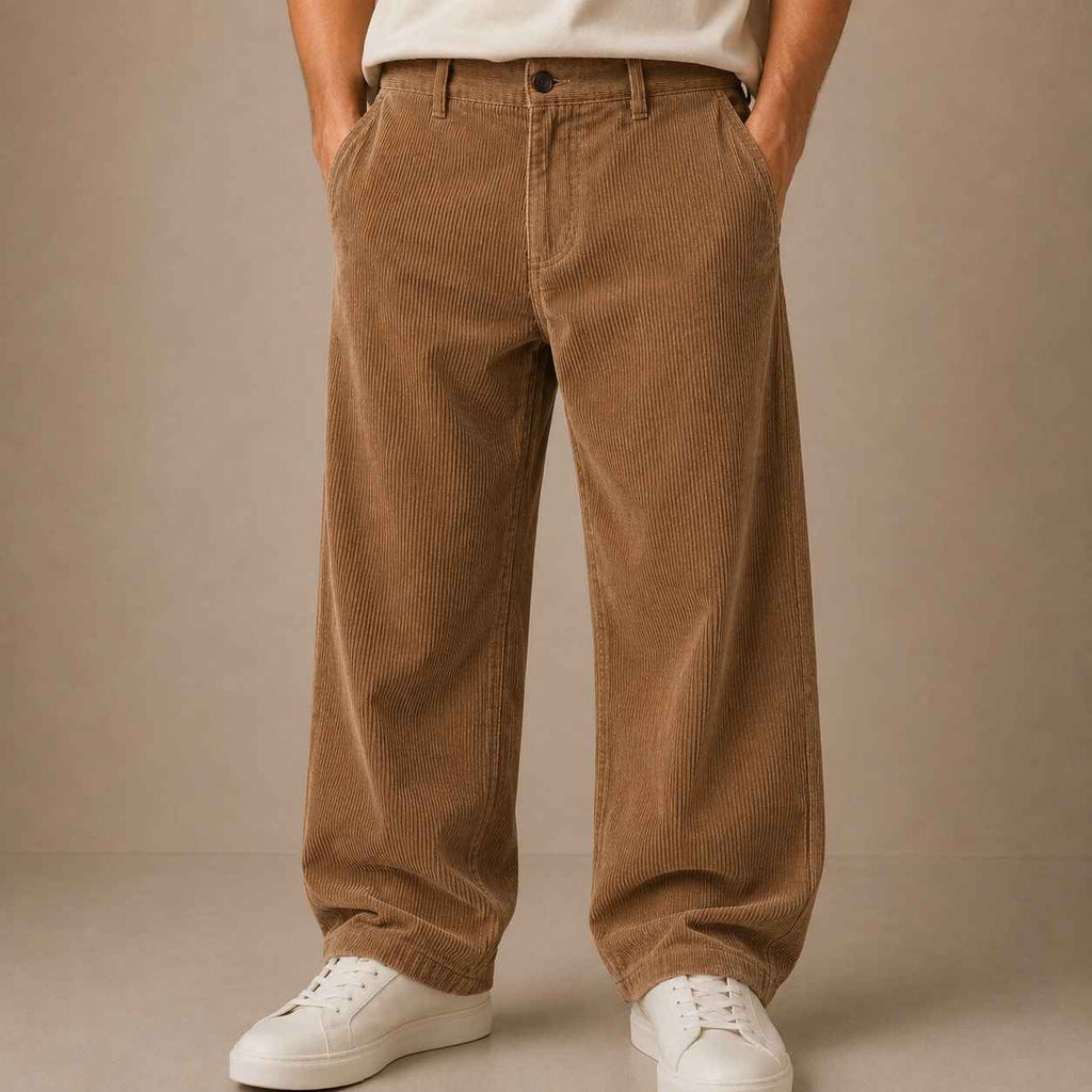 A person wearing loose brown corduroy pants with a white t-shirt and white sneakers, standing against a plain beige studio background.