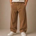 A person wearing loose brown corduroy pants with a white t-shirt and white sneakers, standing against a plain beige studio background.