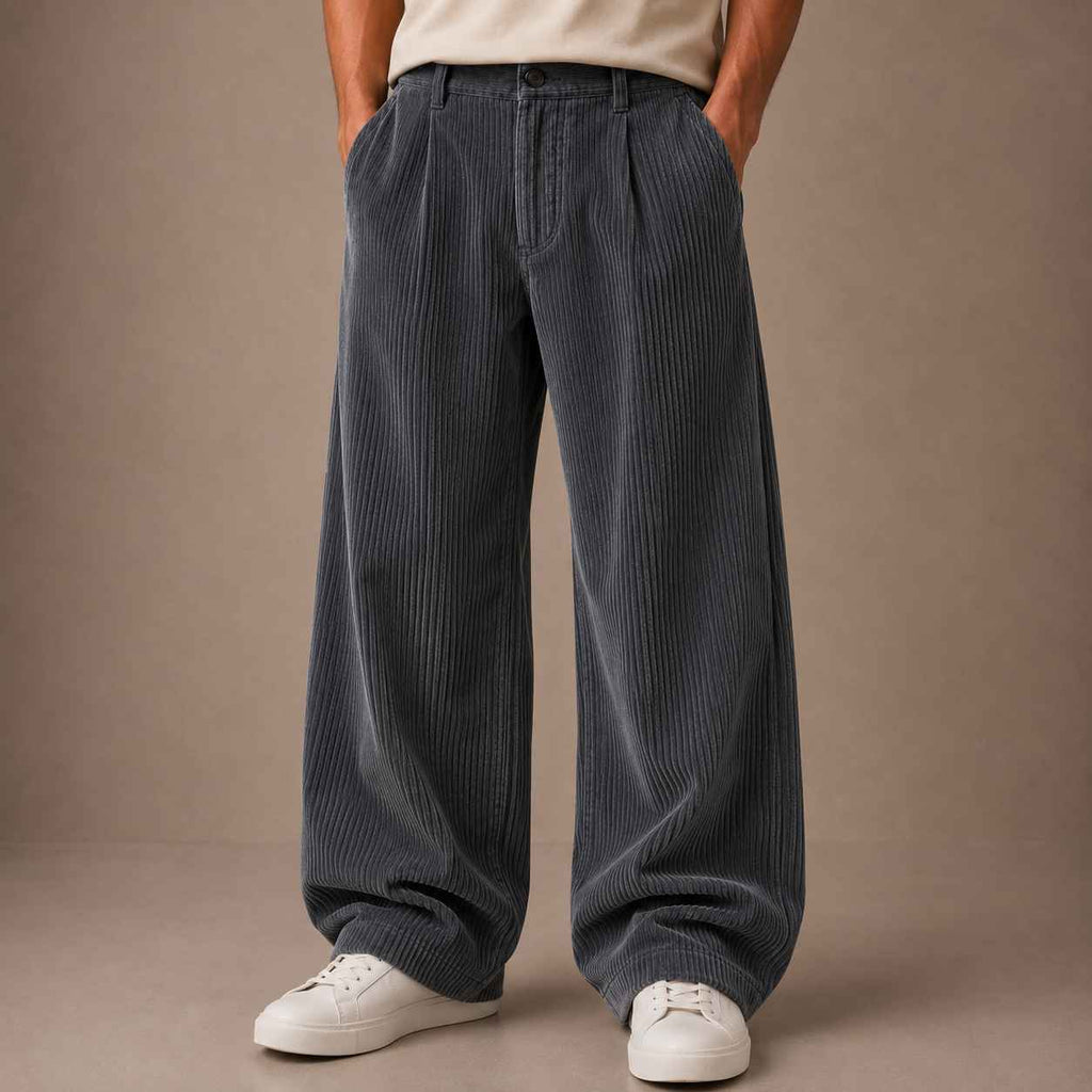 A person wearing wide-leg corduroy trousers in a dark slate gray color, paired with a beige t-shirt and white sneakers, standing against a solid neutral studio backdrop.