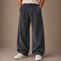 A person wearing wide-leg corduroy trousers in a dark slate gray color, paired with a beige t-shirt and white sneakers, standing against a solid neutral studio backdrop.
