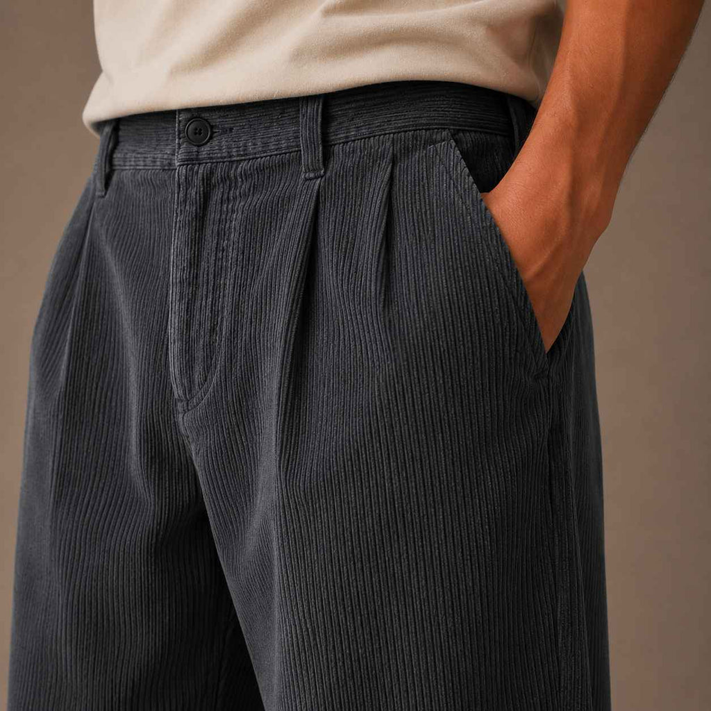 Close-up of a person wearing dark charcoal corduroy trousers with pleats, a beige shirt, and a hand in the pocket, against a warm neutral background.