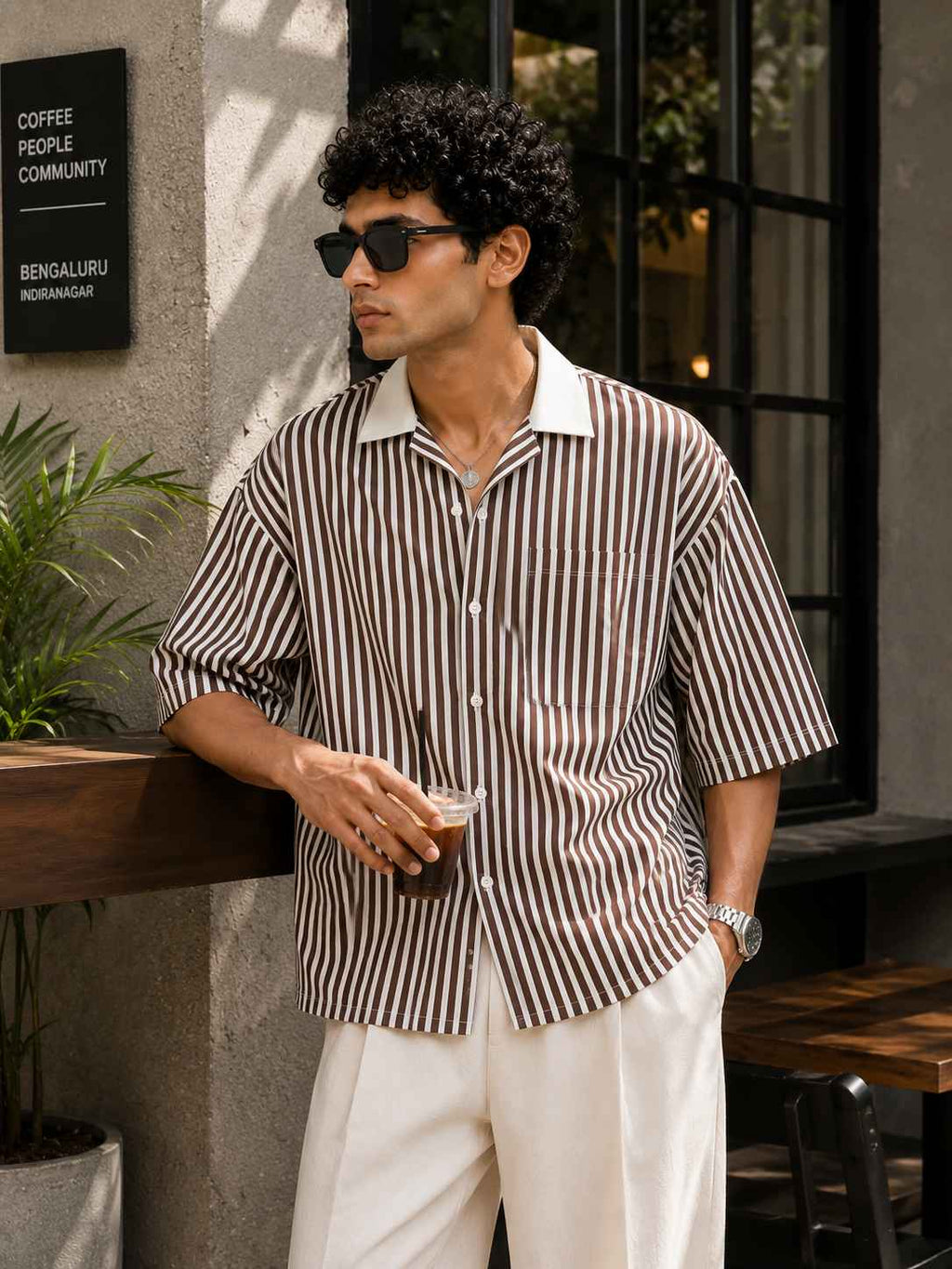 Man in a striped shirt and sunglasses holding a drink outdoors.