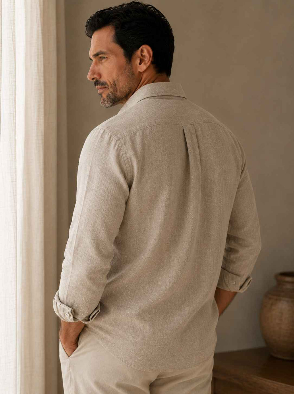 Man wearing a beige shirt and pants standing in a room with a neutral wall and vase.