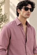 A man wearing a dusty rose linen button-down shirt and black sunglasses, photographed in warm natural light against a light beige wall with soft shadows.