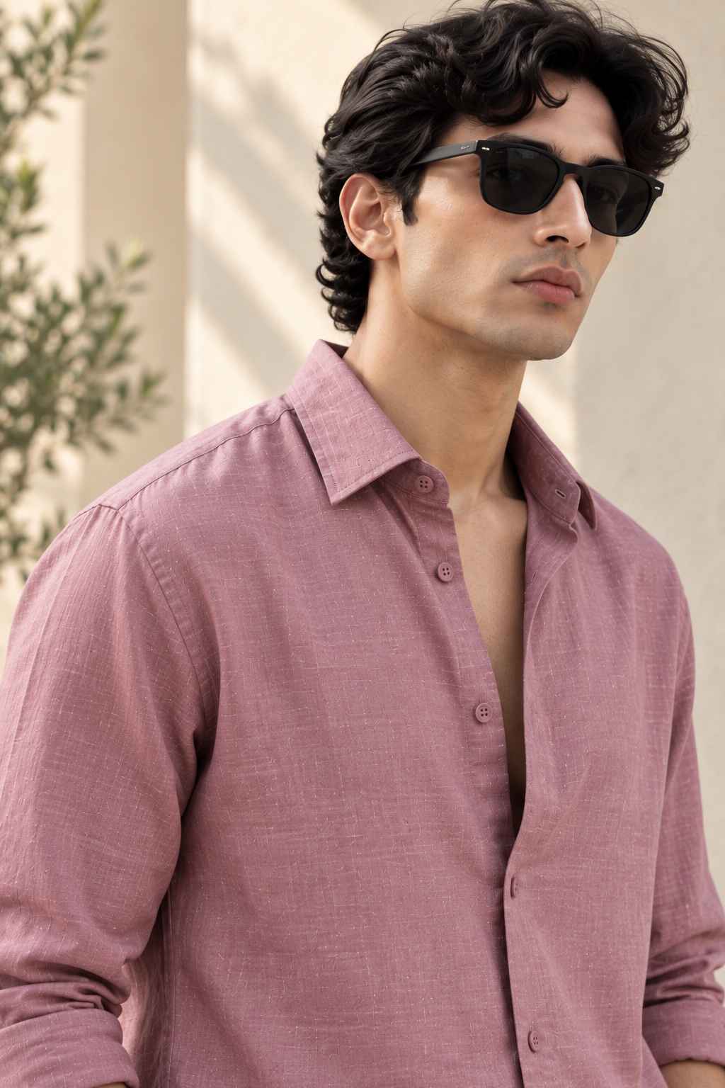 A man wearing a dusty rose linen button-down shirt and black sunglasses, photographed in warm natural light against a light beige wall with soft shadows.