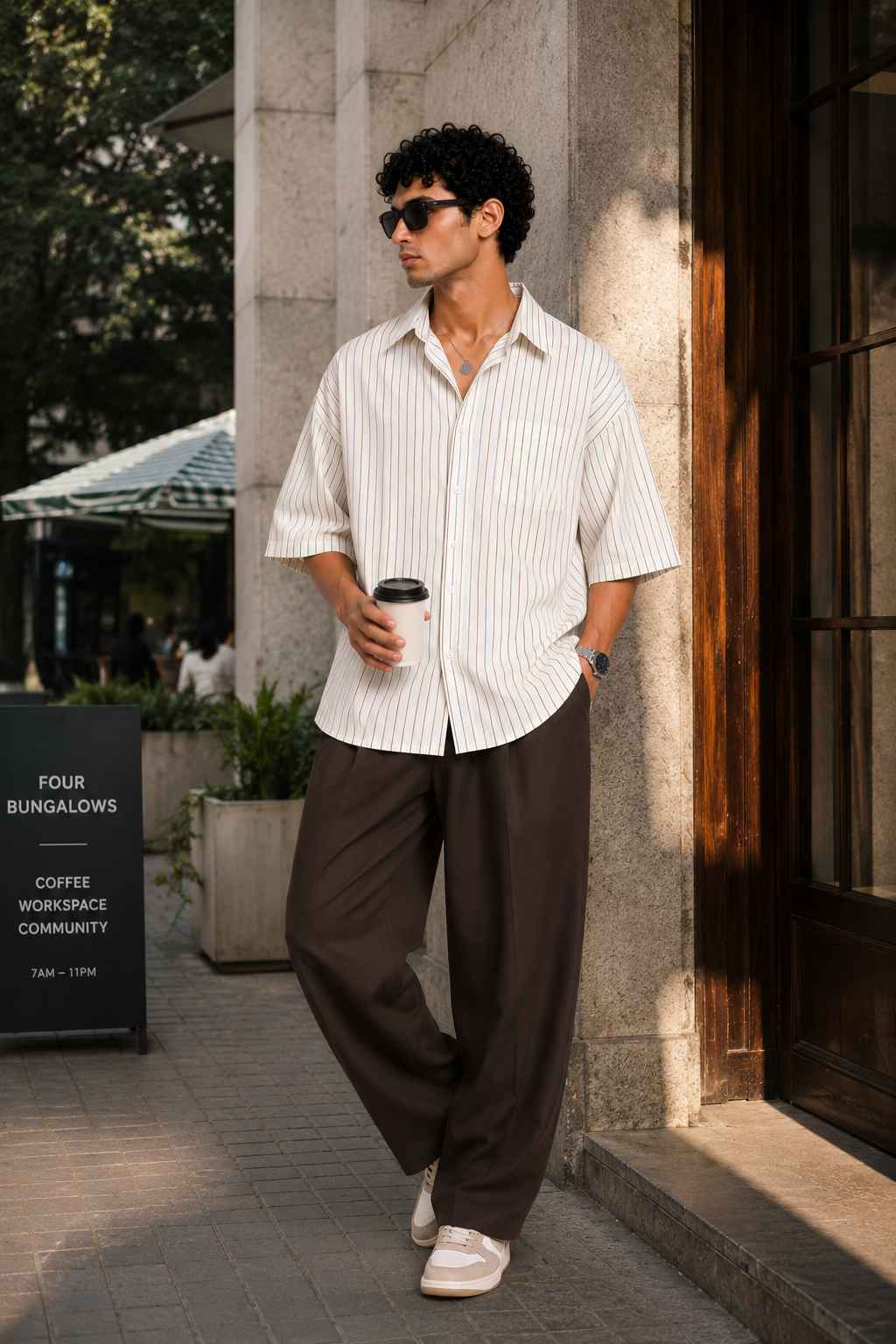 Man walking outdoors holding a coffee cup, wearing a striped shirt and brown pants.