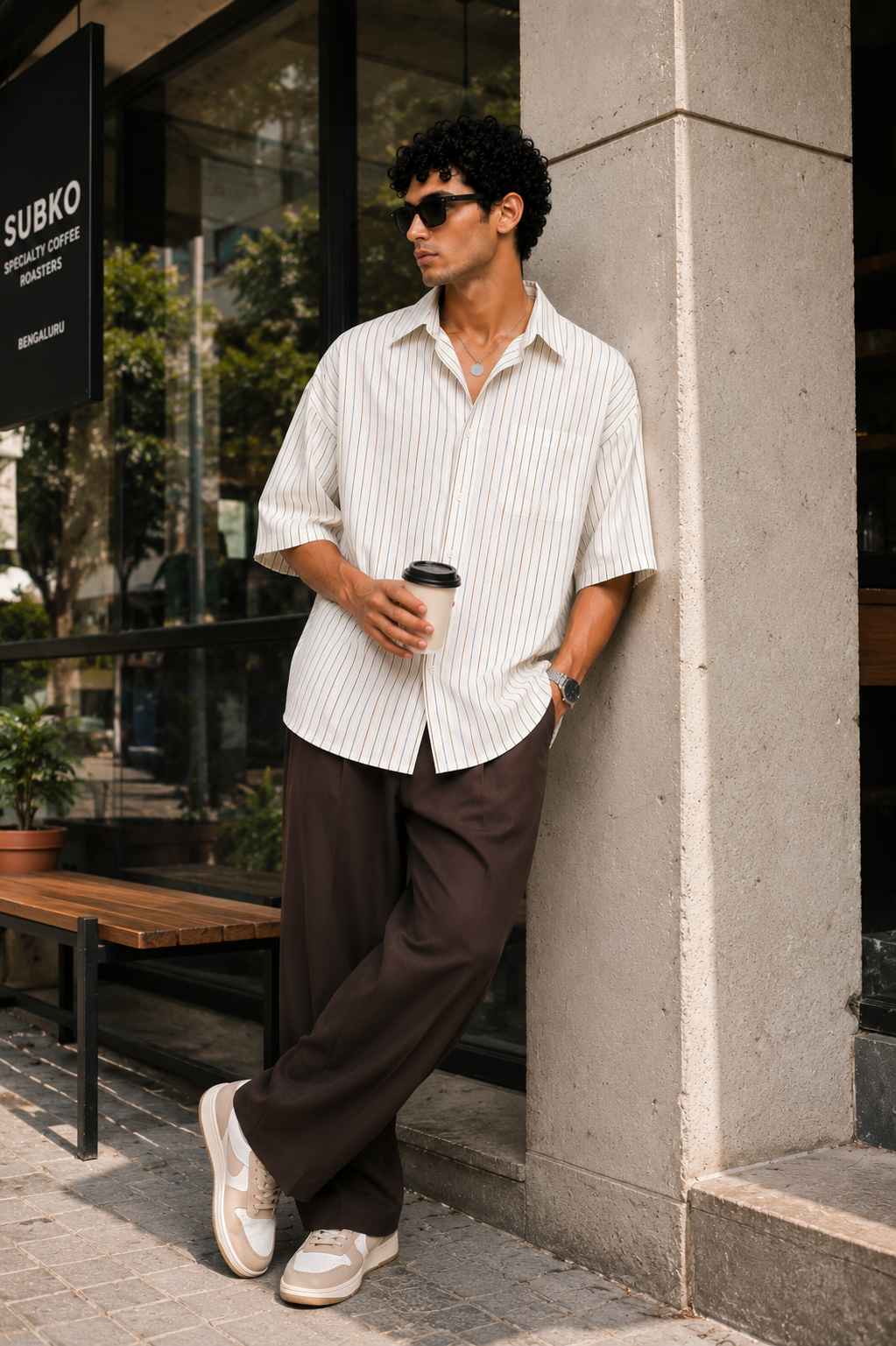 Man leaning against a building holding a coffee cup, wearing sunglasses and a striped shirt.